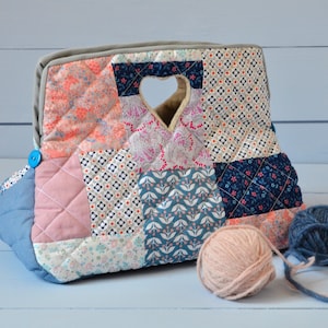 Puede incluir: Una bolsa de tela de patchwork con una abertura en forma de corazón y un botón azul. La bolsa está llena de dos ovillos de lana, uno rosa y otro azul.