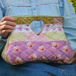 Könnte beinhalten: Gesteppte Patchwork-Handtasche mit herzförmigem Griff. Die Tasche hat verschiedene Blumen- und geometrische Muster in Rosa, Grün und Creme. Die Tasche ist etwa 30 cm breit und 20 cm hoch.