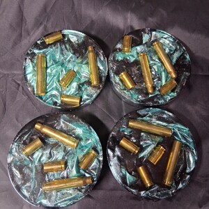 May include: Set of four round coasters. Each coaster features a teal and black resin base with embedded brass bullet casings. The coasters are a unique and decorative item for home decor.