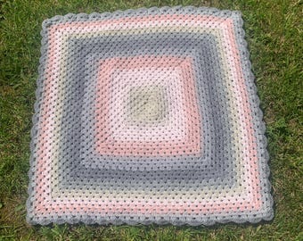 May include: A square, handmade crochet blanket in a granny square pattern. The blanket features concentric squares of pink, gray, and green, with a scalloped edge. The blanket is displayed on a bed of green grass.
