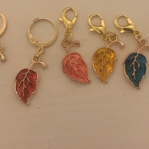 Op de afbeelding: Vijf gouden blaadjes met verschillende geëmailleerde afwerkingen: groen, roze, oranje, geel en blauw. Elke hanger heeft een kleine lus aan de bovenkant om aan een ketting of armband te bevestigen.