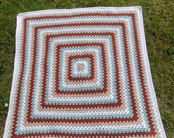 May include: A square, handmade crochet blanket with a concentric square pattern. The blanket features alternating bands of white, light blue, brown, and rust-colored yarn. The blanket is displayed on a bed of green grass.