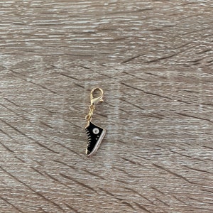 Op de afbeelding: Een gouden hanger in de vorm van een zwarte sneaker met een witte zool en veters. De hanger heeft een kleine gouden lus aan de bovenkant om aan een ketting of armband te bevestigen.
