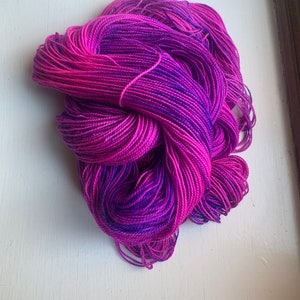 May include: A skein of hand-dyed yarn in shades of pink and purple. The yarn is tightly wound, showing a gradient of colors from bright pink to deep purple. The texture appears soft and suitable for knitting or crochet projects.