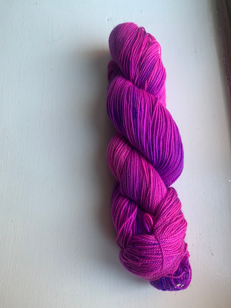 May include: A skein of hand-dyed yarn in shades of pink and purple. The yarn is tightly wound and has a gradient effect, transitioning from a deep magenta to a lighter pink. The yarn is likely made of wool or a similar fiber, suitable for knitting or crochet projects.