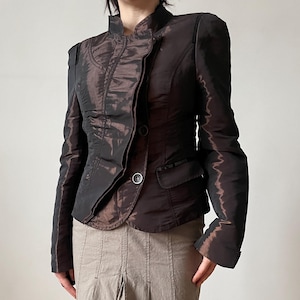 May include: A dark brown, iridescent, structured blazer with a high collar and two buttons, paired with a beige, knee-length skirt featuring vertical paneling, a central inverted pleat, and a dark brown ruffled hem with side drawstring ties.