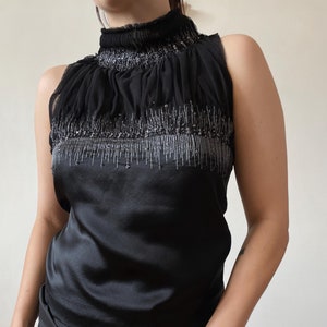 May include: Black sleeveless top with a high, ruffled neckline. The top features a satin bodice and a sheer, embellished upper section with silver beading. The garment is a formal or evening wear piece.