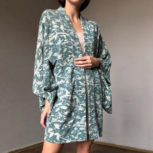 Op de afbeelding: Een blauwgroene jas in kimono-stijl met een all-over wit abstract blad- en takpatroon. Het kledingstuk heeft wijde, losvallende mouwen, een open voorzijde en een rechte zoom die tot halverwege het bovenbeen valt, gemaakt van lichte stof.