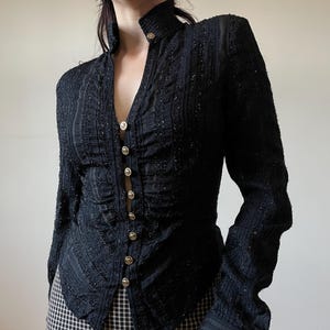 May include: Black long-sleeve jacket with a high collar and gold buttons. The jacket has a textured, embellished design and a fitted silhouette. The buttons run down the front of the jacket. The sleeves have a slight flare at the wrist. The jacket is paired with a black and white checkered skirt.