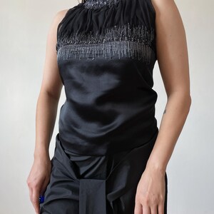 May include: Black sleeveless top with a high, ruched neckline. The top features a satin bodice and is embellished with silver beading along the neckline and chest. The model is wearing black pants. The top is a formal or evening wear garment.