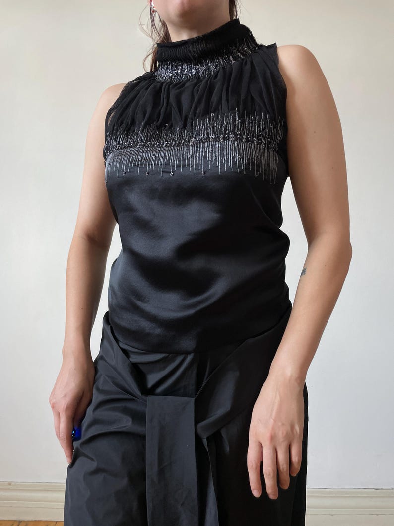May include: Black sleeveless top with a high, ruched neckline. The top features a satin bodice and is embellished with silver beading along the neckline and chest. The model is wearing black pants. The top is a formal or evening wear garment.