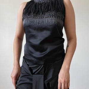 May include: Black sleeveless top with a high, ruffled neckline. The top features a satin bodice and is embellished with silver beading. The matching black skirt has a draped design. The outfit is suitable for formal occasions.