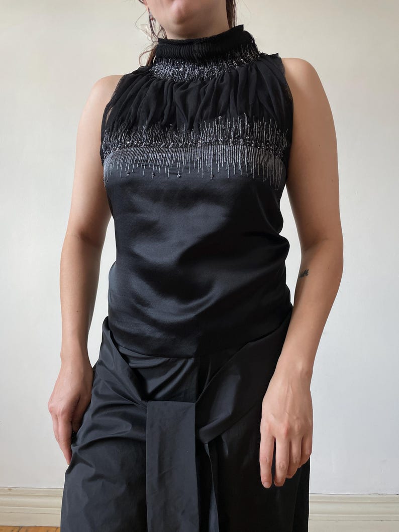 May include: Black sleeveless top with a high, ruffled neckline. The top features a satin bodice and is embellished with silver beading. The matching black skirt has a draped design. The outfit is suitable for formal occasions.