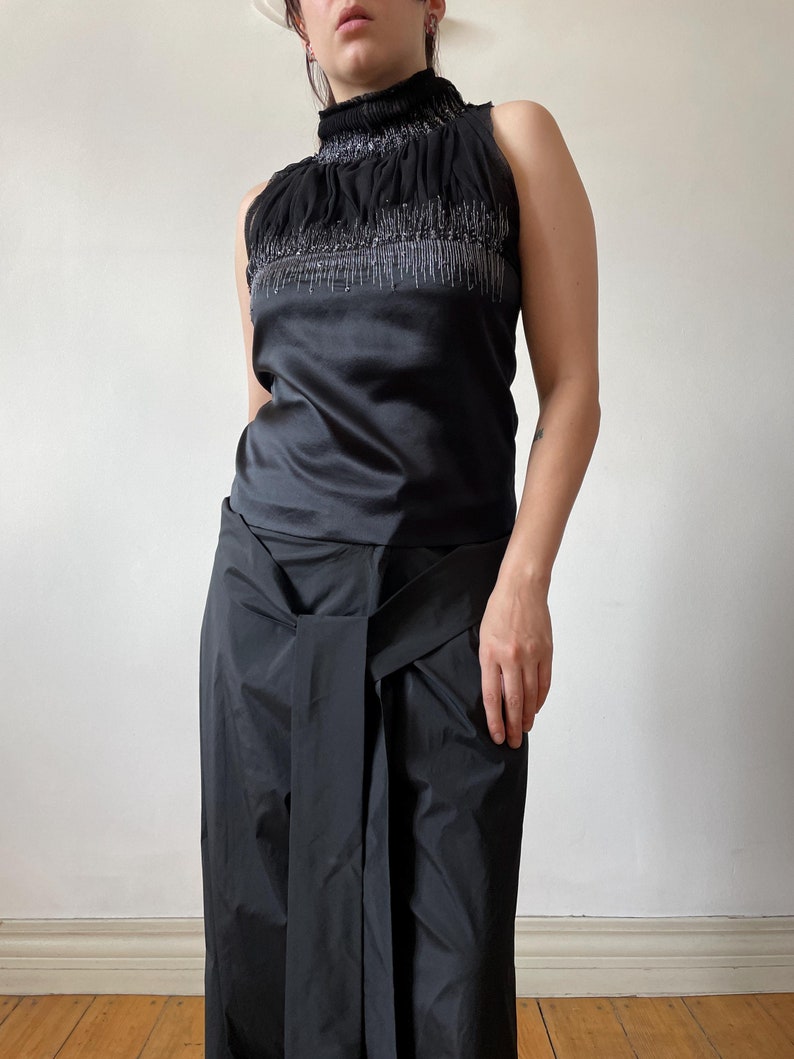 May include: A sleeveless black top with a high, ruffled neckline, embellished with silver beading. The top transitions to a smooth, satin-like fabric. Paired with wide-leg black pants with a wrap-style waist, creating a sleek, monochromatic ensemble.