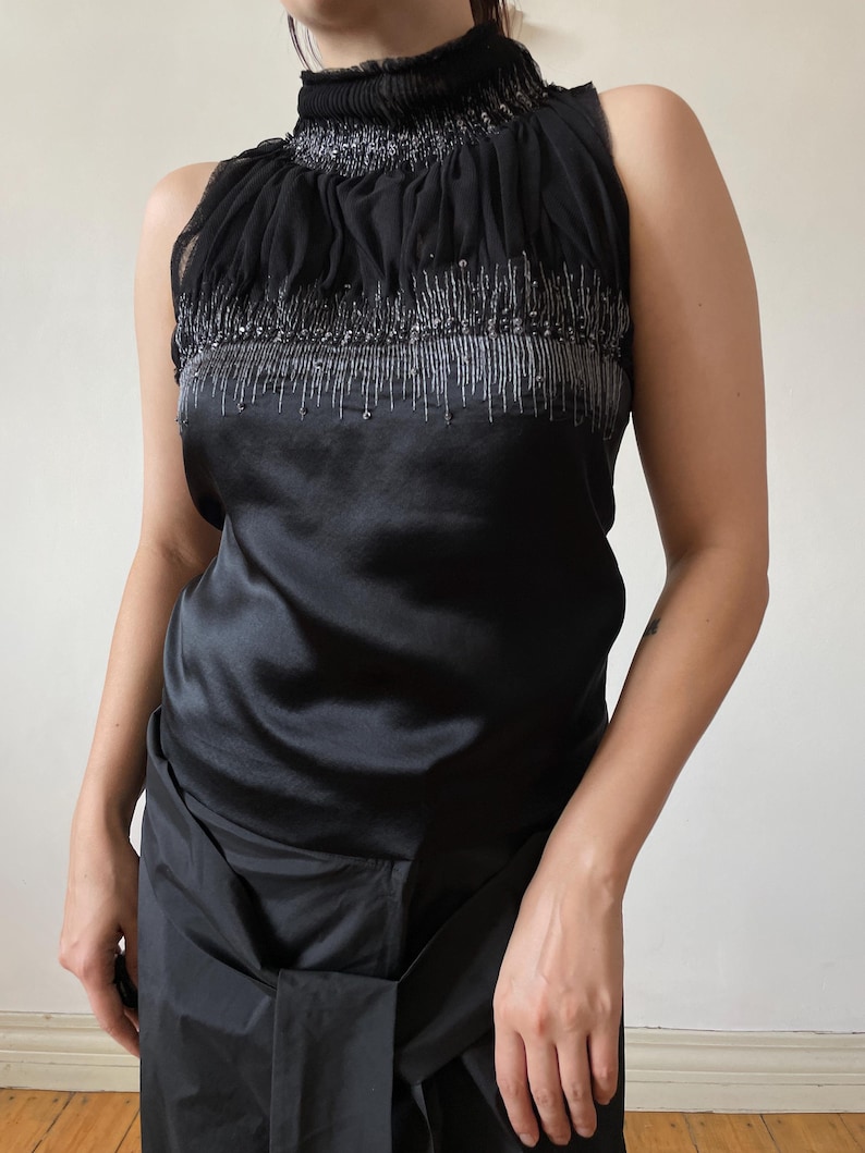 May include: Black sleeveless top with a high, ruffled neckline. The top features a satin bodice and a sheer, embellished upper section with silver beading. The garment is a formal or evening wear piece.