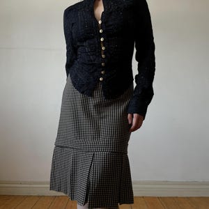 May include: A black, long-sleeved jacket with gold buttons and a high collar paired with a black and white checkered skirt. The jacket has a textured appearance. The skirt has a pleated hemline.