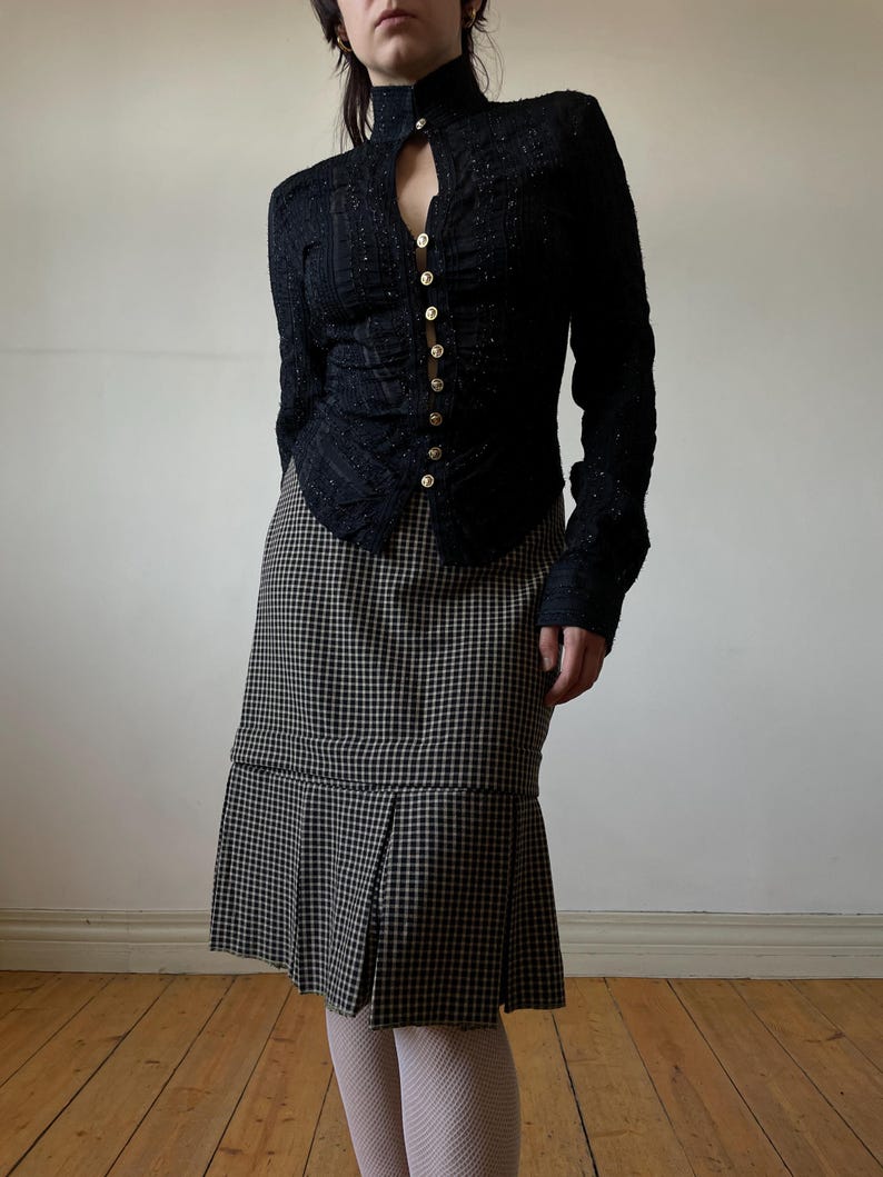 May include: A black, long-sleeved jacket with gold buttons and a high collar paired with a black and white checkered skirt. The jacket has a textured appearance. The skirt has a pleated hemline.