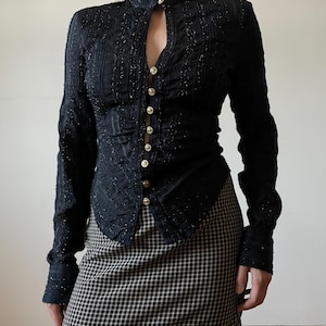 May include: Black long-sleeve jacket with a high collar and gold buttons. The jacket has a textured, embellished design and a keyhole neckline. The model is wearing a black and white checkered skirt.