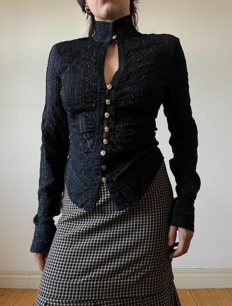 May include: Black long-sleeve jacket with a high collar and gold buttons. The jacket has a textured, embellished design and a keyhole neckline. The model is wearing a black and white checkered skirt.