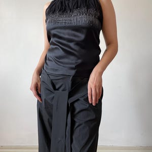 May include: Sleeveless black top with a high, ruffled neckline and silver beaded embellishments. The top has a satin finish and is paired with matching black, wide-leg pants with a wrap-around waist detail. The outfit is a sleek, monochromatic ensemble.