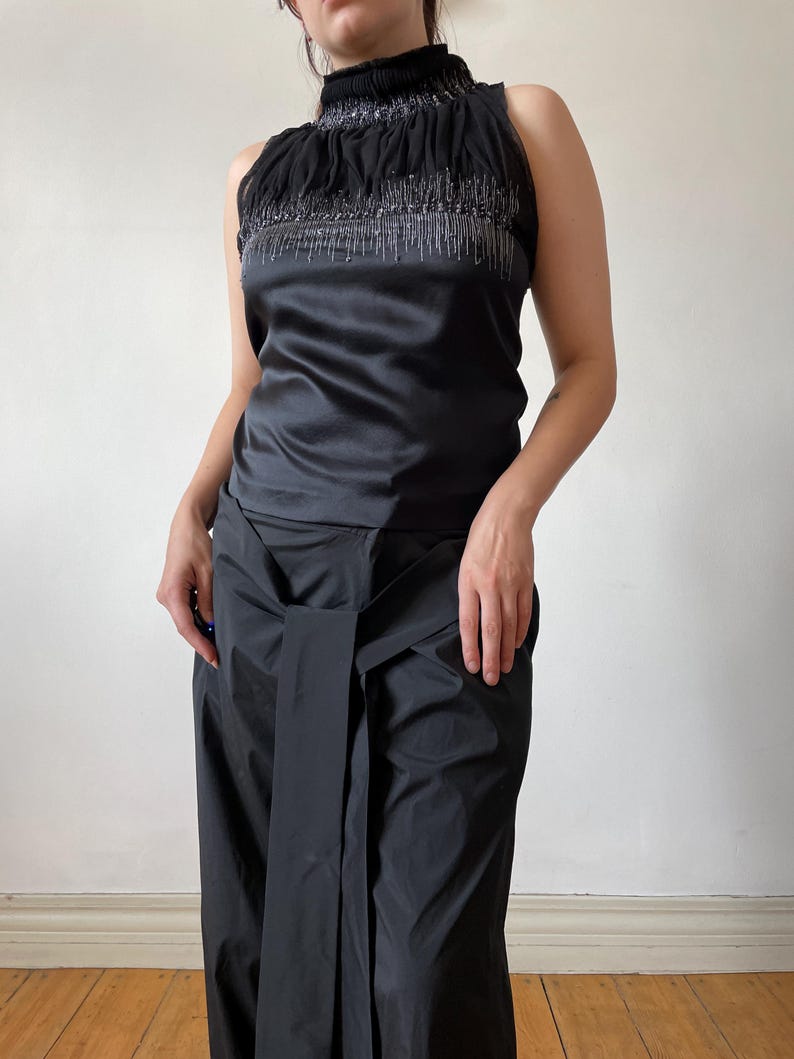 May include: Sleeveless black top with a high, ruffled neckline and silver beaded embellishments. The top has a satin finish and is paired with matching black, wide-leg pants with a wrap-around waist detail. The outfit is a sleek, monochromatic ensemble.