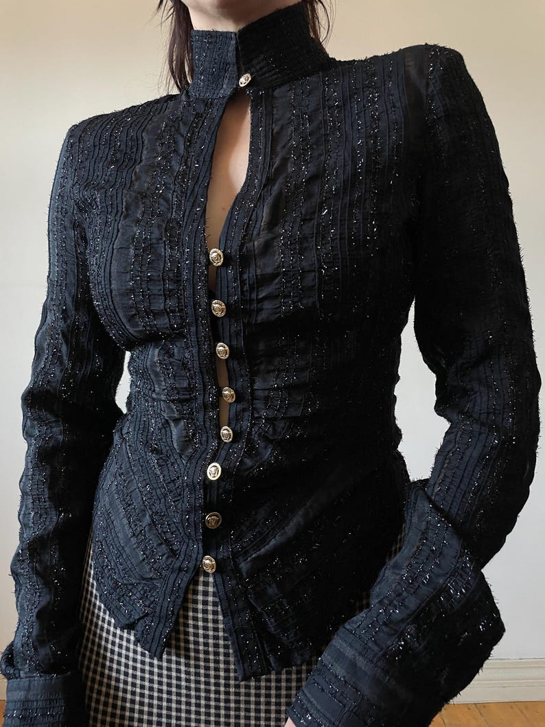 May include: Black long-sleeve blouse with a high collar and gold buttons. The blouse features vertical textured stripes and a slight sparkle throughout the fabric. The sleeves have a flared design at the wrist, and the blouse is buttoned down the front.