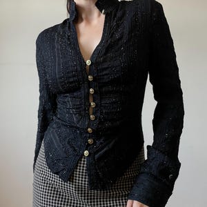 May include: Black long-sleeve button-down top with a high collar and gold buttons. The top has a textured, shimmering fabric and is paired with a black and white checkered skirt. The top's design includes vertical stripes and a V-neck.