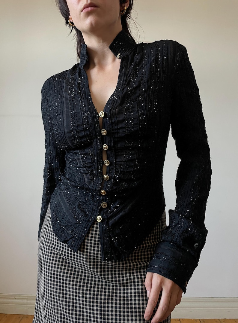 May include: Black long-sleeve button-down top with a high collar and gold buttons. The top has a textured, shimmering fabric and is paired with a black and white checkered skirt. The top's design includes vertical stripes and a V-neck.