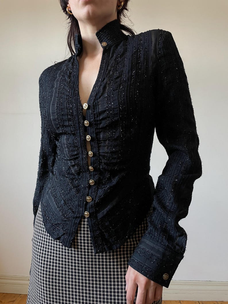 May include: Black long-sleeve jacket with a high collar and gold buttons. The jacket has a textured, embellished design and a fitted silhouette. The buttons run down the front of the jacket. The sleeves have a slight flare at the wrist. The jacket is paired with a black and white checkered skirt.