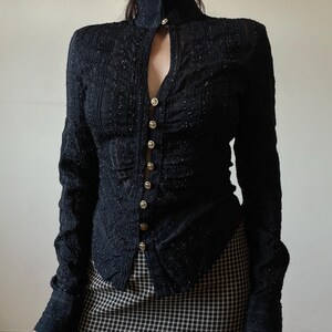 May include: Black long-sleeved top with a high collar and keyhole neckline. The top features vertical textured detailing and is fastened with gold-colored buttons. The sleeves have textured cuffs. The top is paired with a black and white checkered skirt.