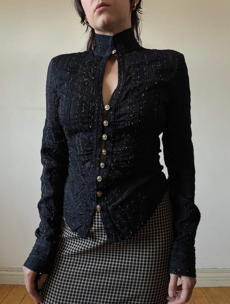 May include: Black long-sleeved top with a high collar and keyhole neckline. The top features vertical textured detailing and is fastened with gold-colored buttons. The sleeves have textured cuffs. The top is paired with a black and white checkered skirt.