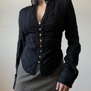 May include: Black long-sleeve jacket with gold buttons. The jacket has a high collar and a textured, sparkly fabric. The buttons run down the front. The model is wearing a black and white checkered skirt.