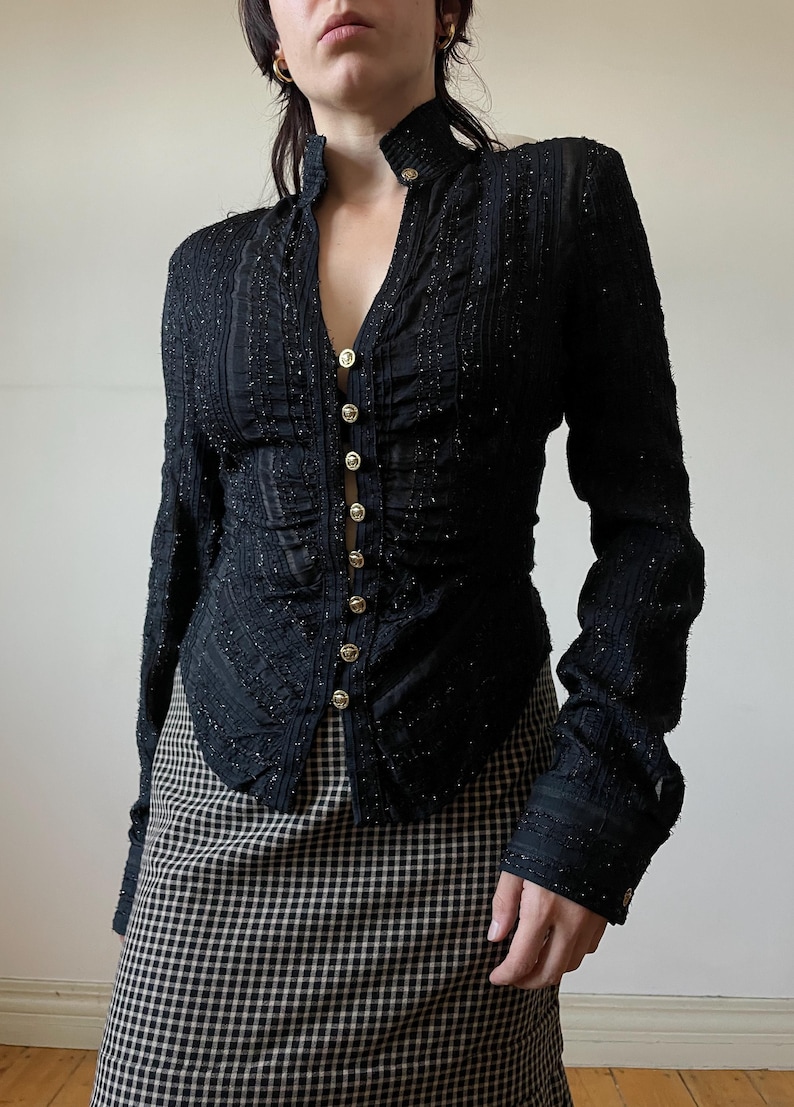 May include: Black long-sleeve jacket with gold buttons. The jacket has a high collar and a textured, sparkly fabric. The buttons run down the front. The model is wearing a black and white checkered skirt.