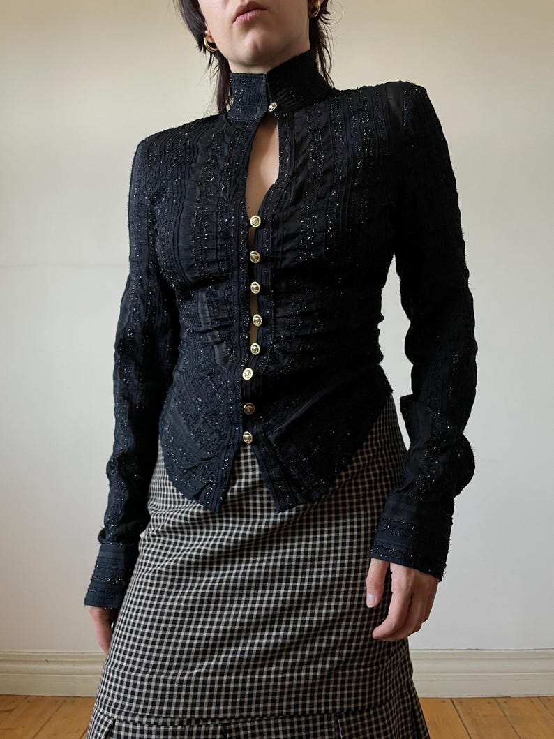 May include: Black long-sleeve top with a high collar and gold buttons. The top has a textured, embellished design and a keyhole detail at the neckline. The model is wearing a black and white plaid skirt.