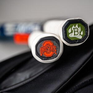 May include: Two white tennis rackets with black and green end caps. The end caps have logos on them. One logo is a paw print with the letter "B" inside. The other logo is the Ohio State University logo.