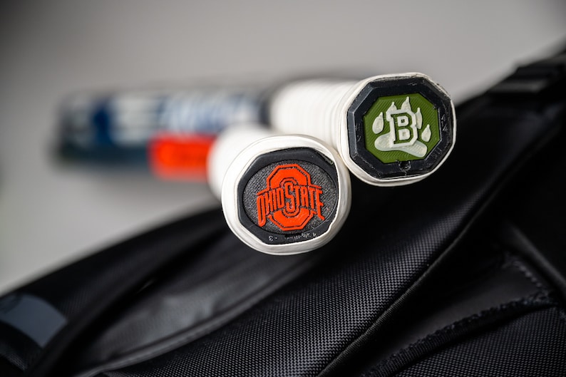 May include: Two white tennis rackets with black and green end caps. The end caps have logos on them. One logo is a paw print with the letter "B" inside. The other logo is the Ohio State University logo.