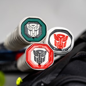 May include: Three tennis racket grip caps with a silver base and a red, green, or white octagon with a black Transformers logo inside.