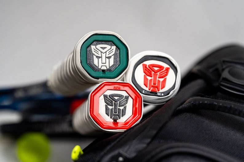 May include: Three tennis racket grip caps with a silver base and a red, green, or white octagon with a black Transformers logo inside.