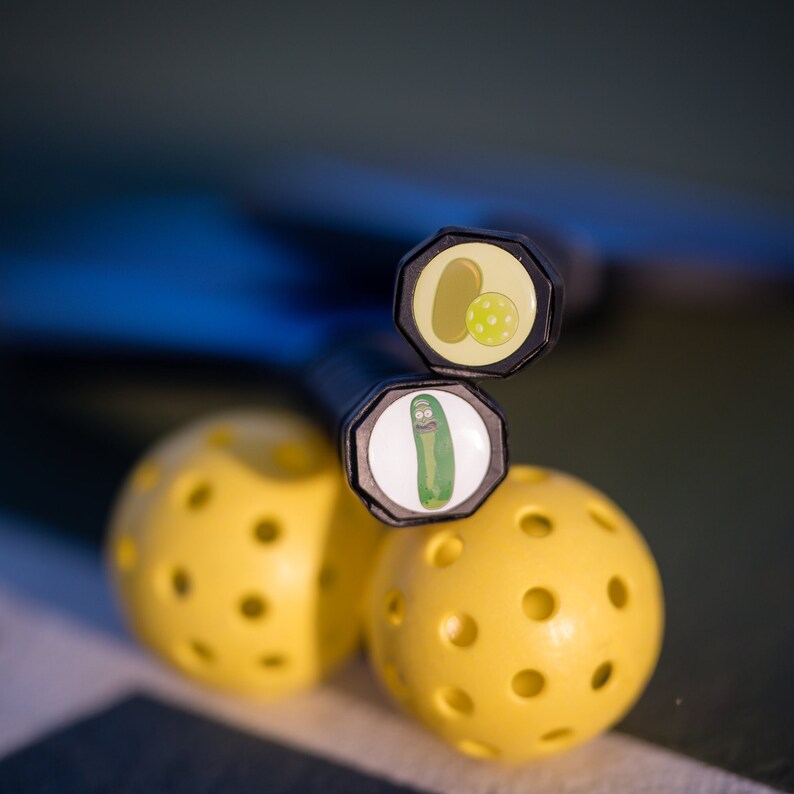 May include: Two yellow pickleball paddles with black handles. The handles have round black caps with cartoon images of a pickle and a pickle character.