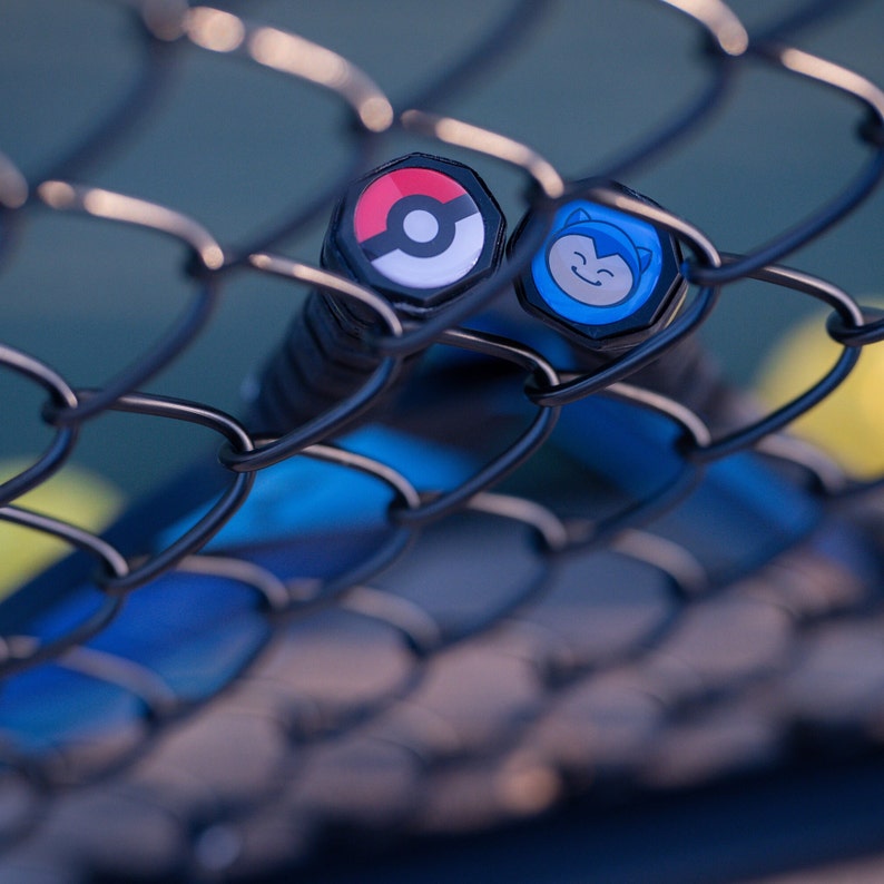May include: Two tennis rackets with blue and black grips, seen through a chain link fence. The ends of the rackets have colorful cartoon character designs. One design is a red and white Pokeball, the other is a blue cartoon character with a white face.