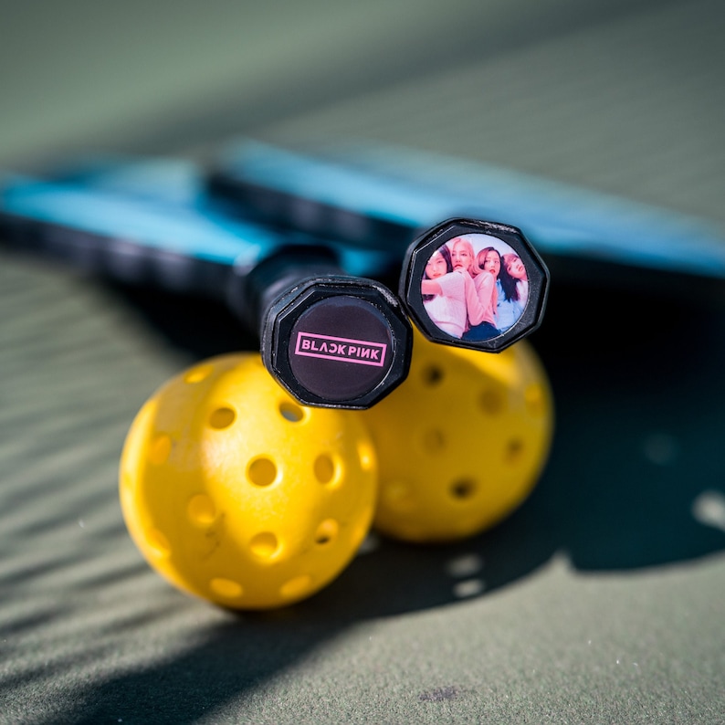 May include: Two yellow pickleball paddles with black handles. One handle has a black and white sticker with the text "BLACKPINK" in pink letters. The other handle has a sticker with a photo of a group of women.