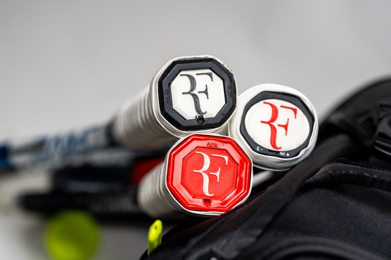 May include: Three tennis rackets with black, white, and red grips featuring the "RF" logo. The rackets are partially visible in a black tennis bag.