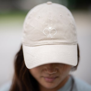 May include: A beige baseball cap with a white embroidered design of two pickleball paddles and a ball.