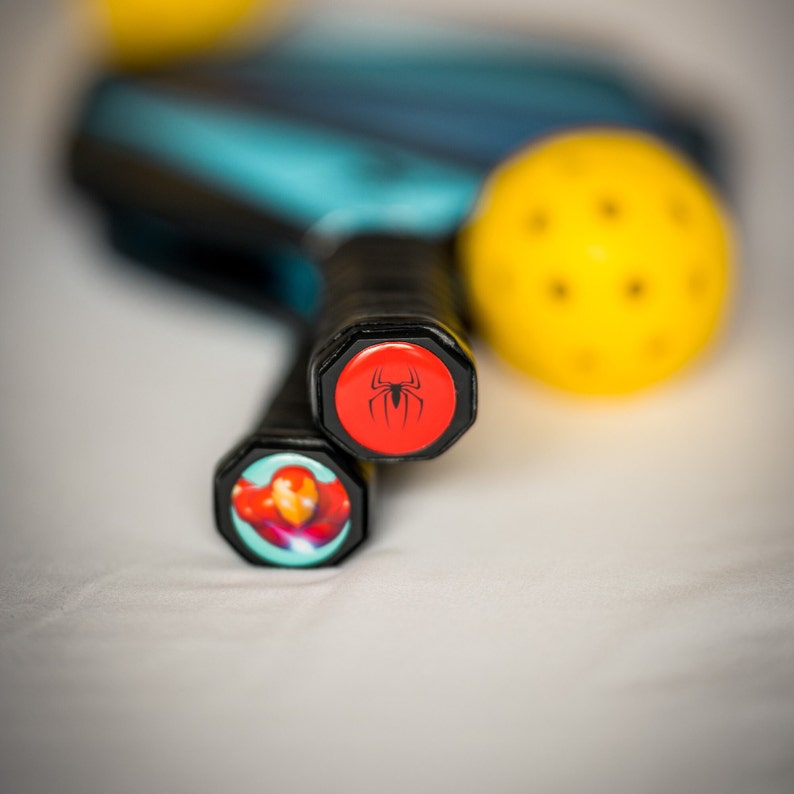 May include: Two black pickleball paddles with red and blue accents. The top paddle has a red cap with a black spider logo. The bottom paddle has a blue cap with a red and yellow Iron Man logo.