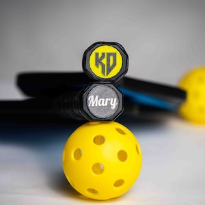 May include: A yellow pickleball with black holes sits on top of two black pickleball paddles. The top paddle has a yellow cap with the initials "KD" in black. The bottom paddle has a gray cap with the name "Mary" in white.