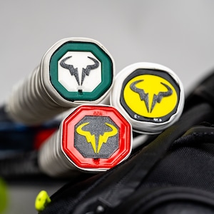 May include: Three tennis rackets with different colored grip ends. The ends are octagonal and feature a bull logo with a lightning bolt. The colors are green, yellow, and red.