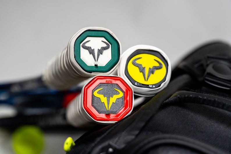 May include: Three tennis rackets with different colored grip ends. The ends are octagonal and feature a bull logo with a lightning bolt. The colors are green, yellow, and red.