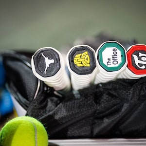 May include: Four tennis racket grips with different logos. The logos are a longhorn steer, a transformer, "The Office", and a "T" with a swirl.