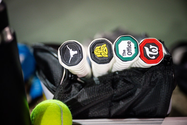 May include: Four tennis racket grips with different logos. The logos are a longhorn steer, a transformer, "The Office", and a "T" with a swirl.