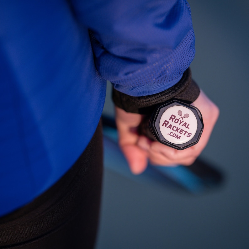 May include: A person is holding a black tennis racket with a white sticker on the end. The sticker says "Royal Rackets .com" and has a tennis racket icon.